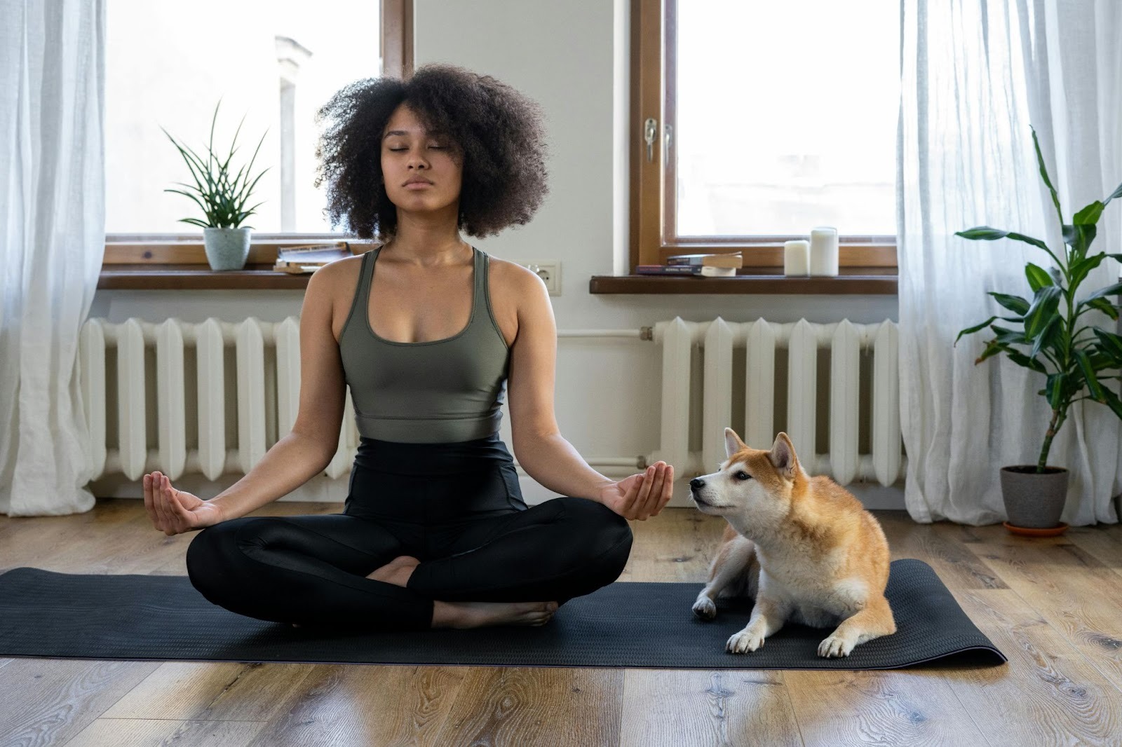 Mulher negra praticando meditação com o seu cachorro ao lado. Ela está sentada em um tapete de exercícios, com as pernas cruzadas, em posição de concentração.