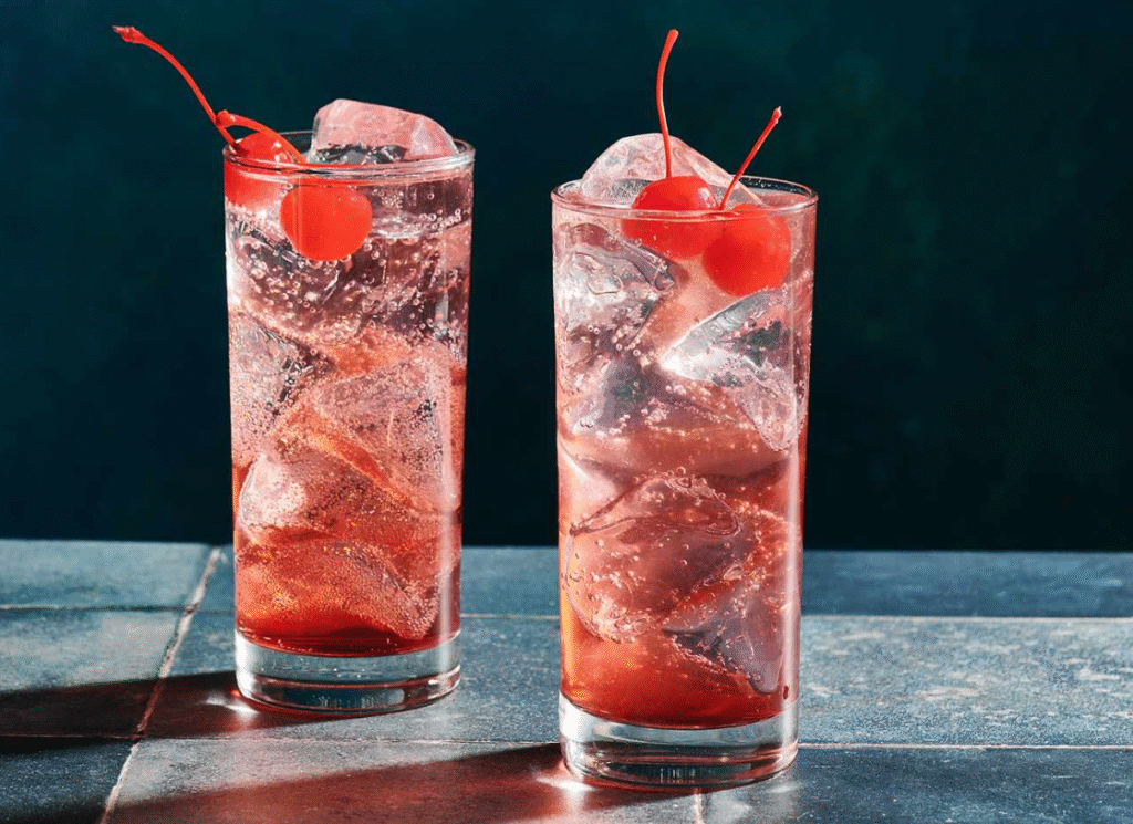 Dois copos altos de Shirley Temple, drink sem álcool com gelo, refrigerante de limão e grenadine, decorados com cerejas ao marasquino.